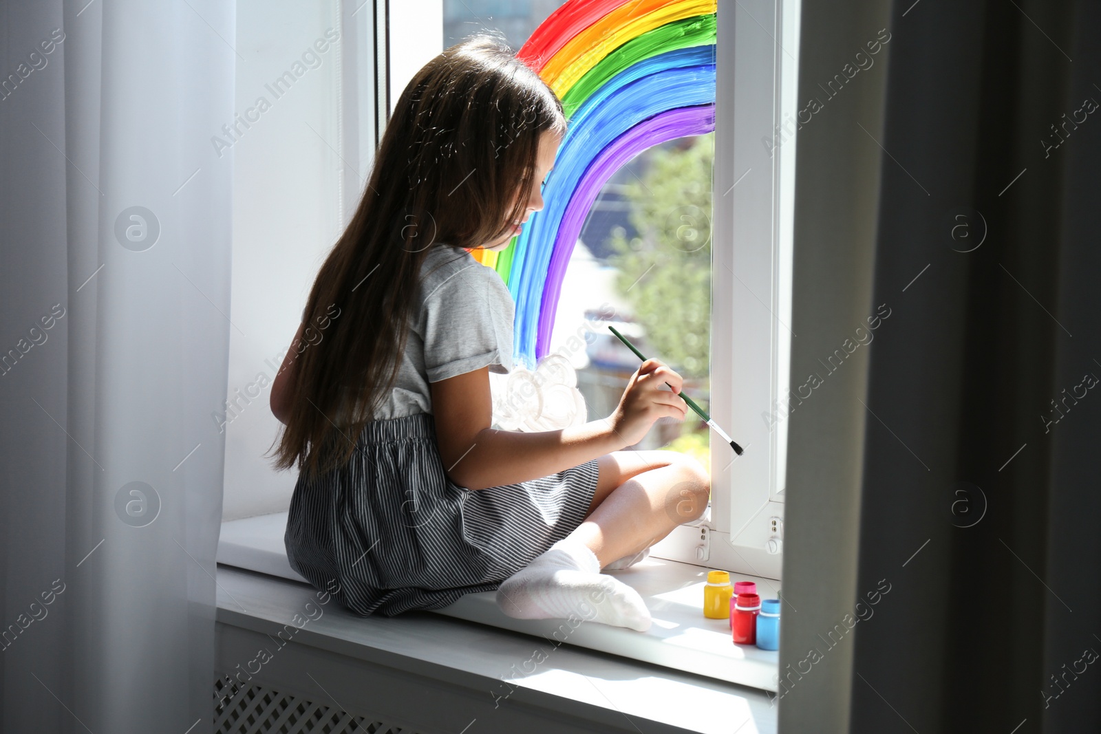 Photo of Little girl drawing rainbow on window indoors. Stay at home concept