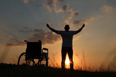 Photo of Man raising hands up to sky near wheelchair at sunset, back view. Healing miracle