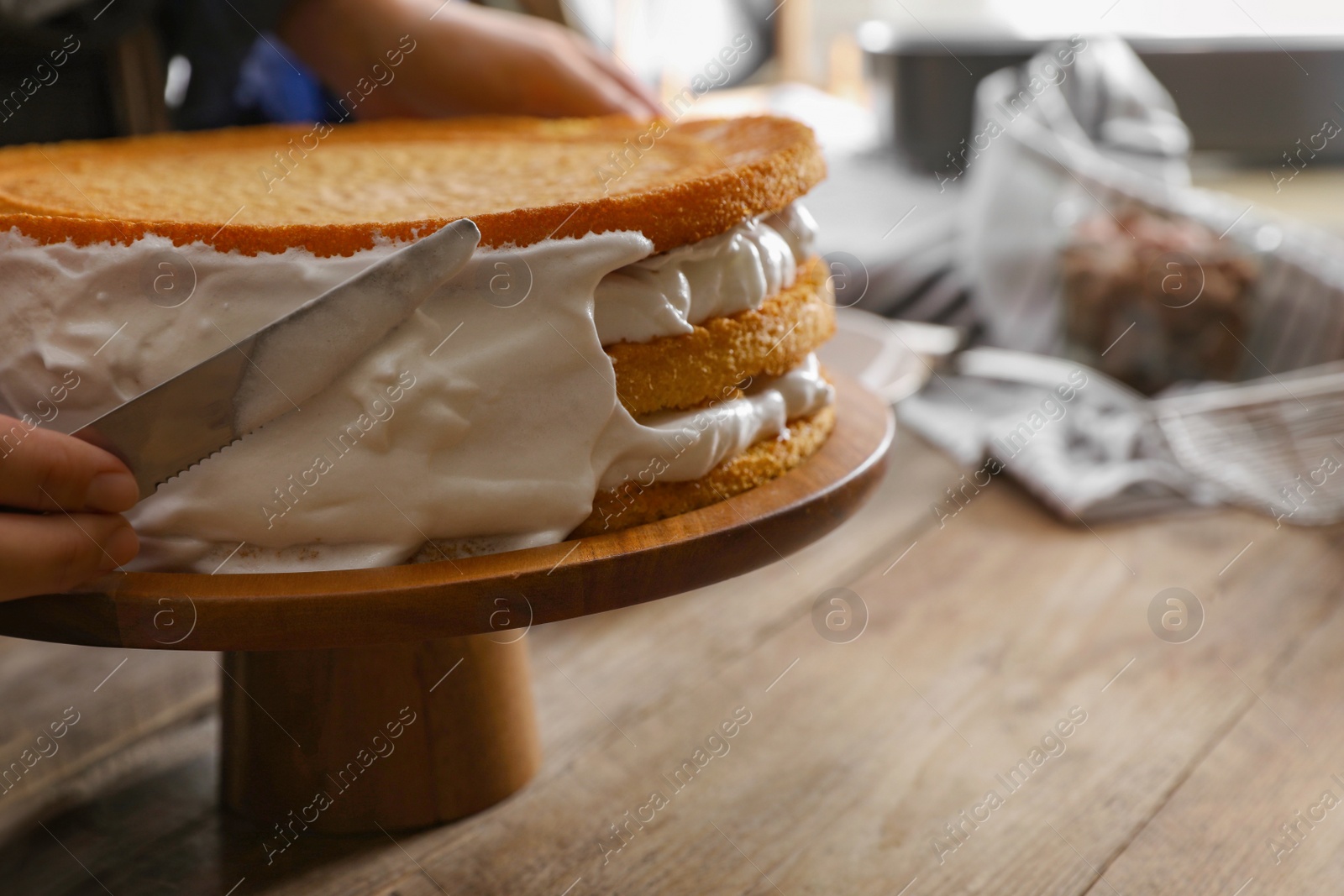 Photo of Woman smearing sides of sponge cake with cream at wooden table, closeup
