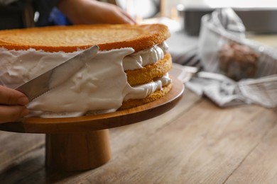 Woman smearing sides of sponge cake with cream at wooden table, closeup