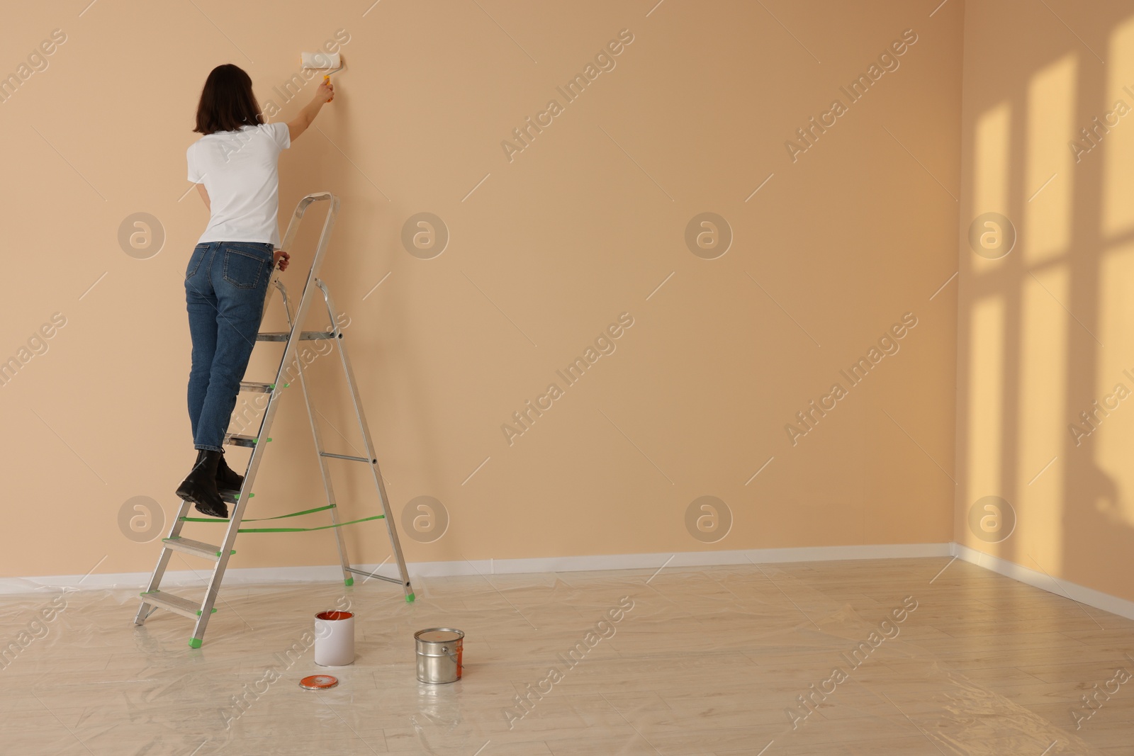 Photo of Young woman painting wall with roller on stepladder indoors, space for text. Room renovation