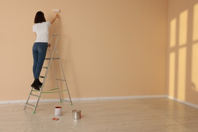 Young woman painting wall with roller on stepladder indoors, space for text. Room renovation