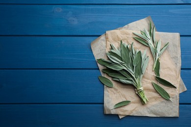 Photo of Bunch of fresh green sage with parchment on blue wooden table, flat lay. Space for text