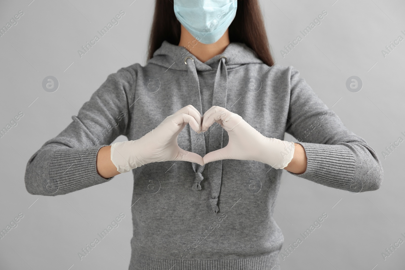 Photo of Woman in protective face mask and medical gloves making heart with hands on grey background, closeup