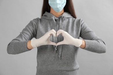 Photo of Woman in protective face mask and medical gloves making heart with hands on grey background, closeup