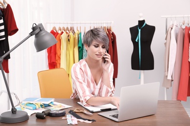 Photo of Beautiful young stylist talking on phone at workplace near rack with clothes