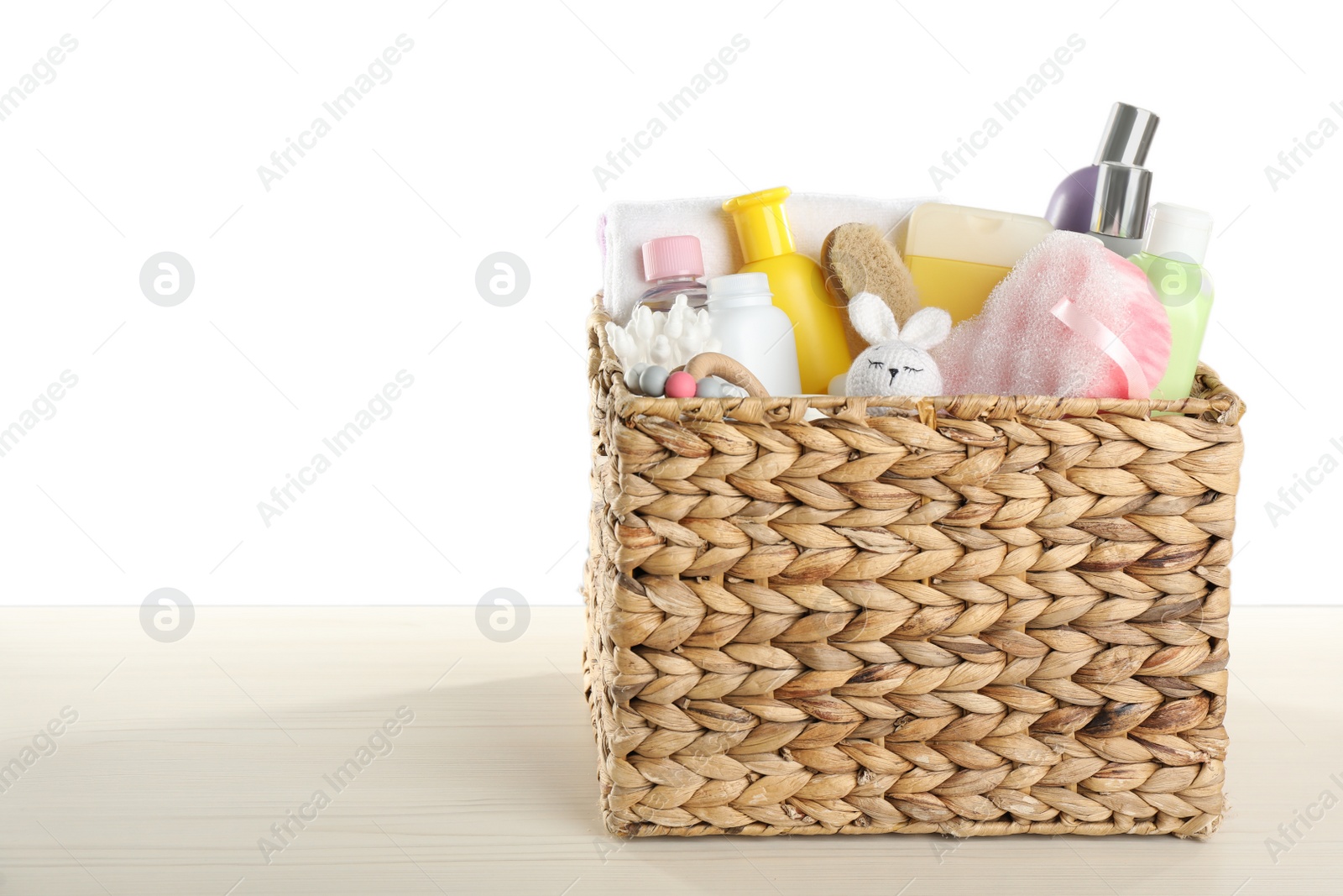 Photo of Wicker basket full of different baby cosmetic products, bathing accessories and toys on wooden table against white background