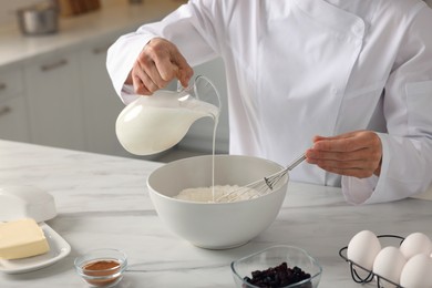 Photo of Professional chef adding milk into dough at white marble table indoors, closeup