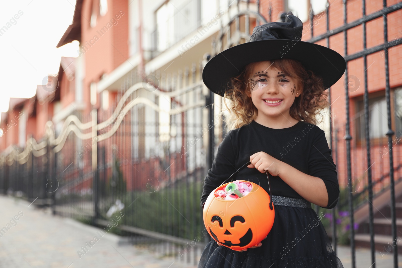 Photo of Cute little girl with pumpkin candy bucket wearing Halloween costume outdoors