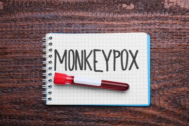 Photo of Word Monkeypox written in notebook and test tube with blood sample on wooden table, top view