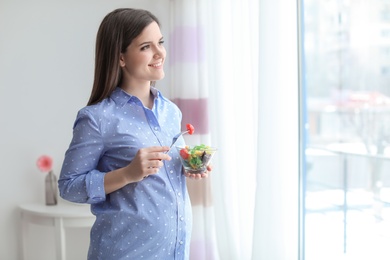 Photo of Young pregnant woman eating vegetable salad near window at home