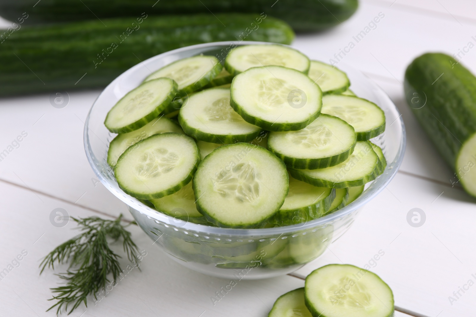 Photo of Cut cucumber in glass bowl, fresh vegetables and dill on white wooden table, closeup