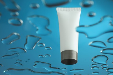 Photo of Moisturizing cream in tube on glass with water drops against blue background, low angle view. Space for text