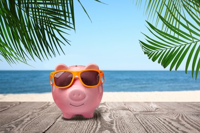 Image of Saving money for summer vacation. Piggy bank with sunglasses on wooden surface near sandy beach and sea