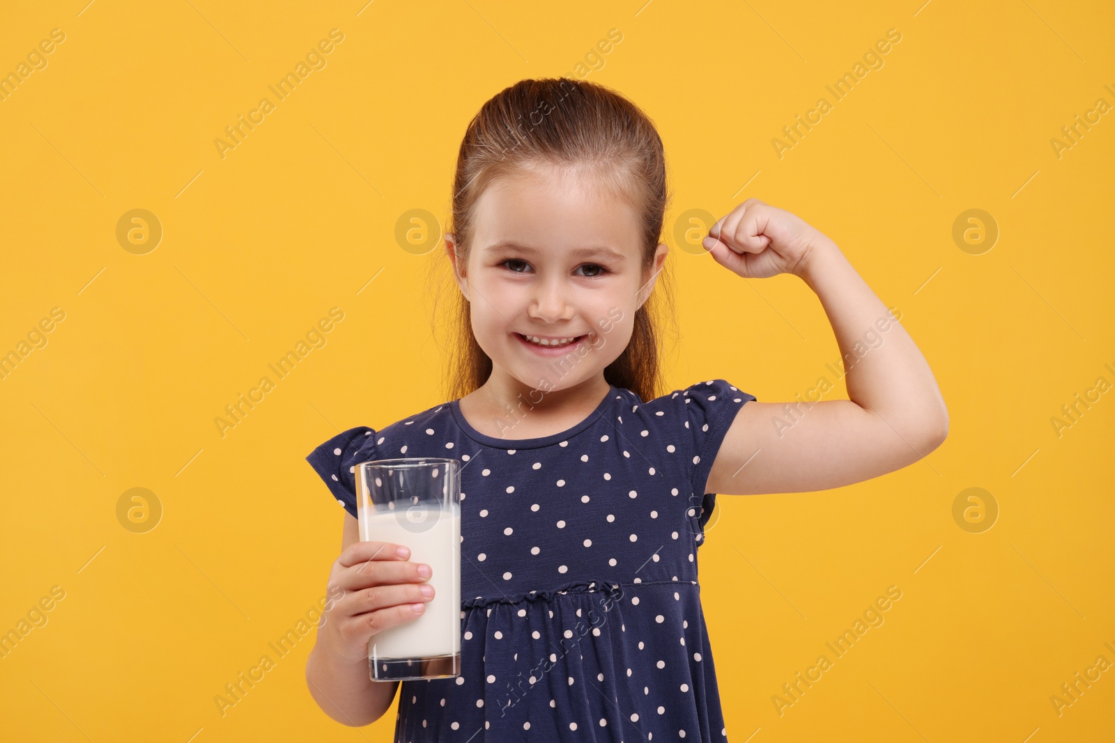 Photo of Cute girl with glass of fresh milk showing her strength on orange background