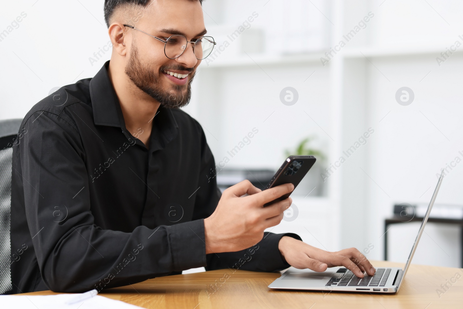 Photo of Handsome young man using smartphone while working with laptop at wooden table in office
