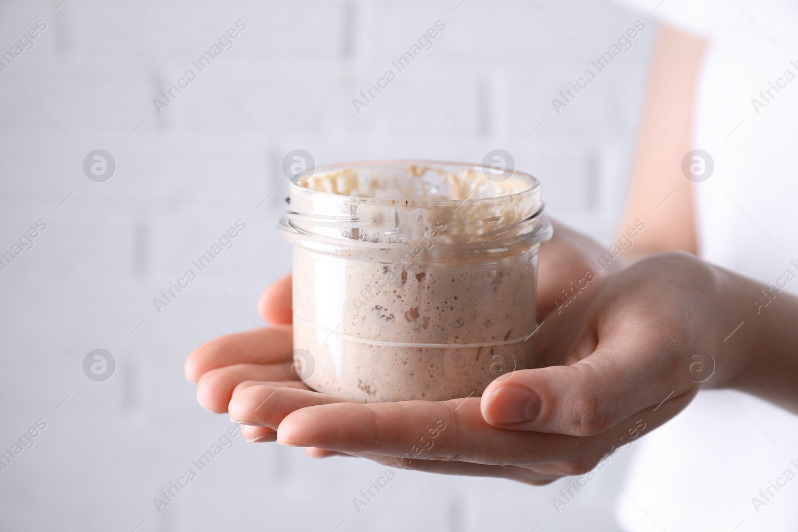 Photo of Woman holding glass jar with fresh sourdough starter on blurred background, closeup. Space for text