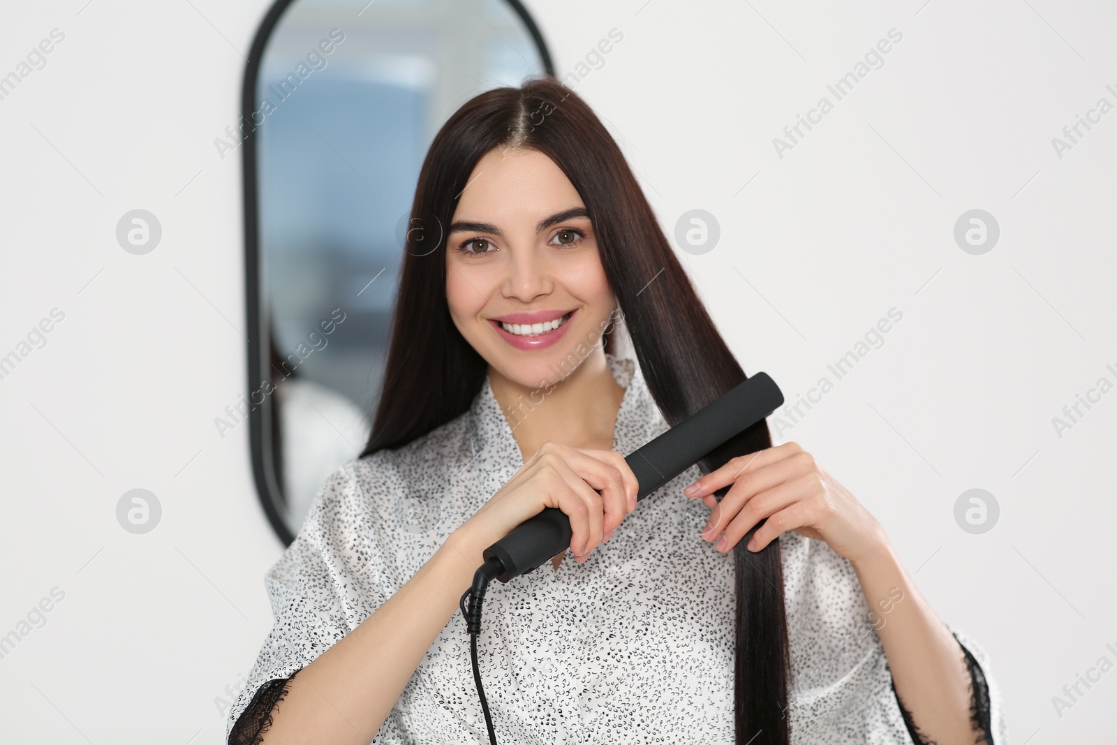 Photo of Beautiful happy woman using hair iron near mirror indoors