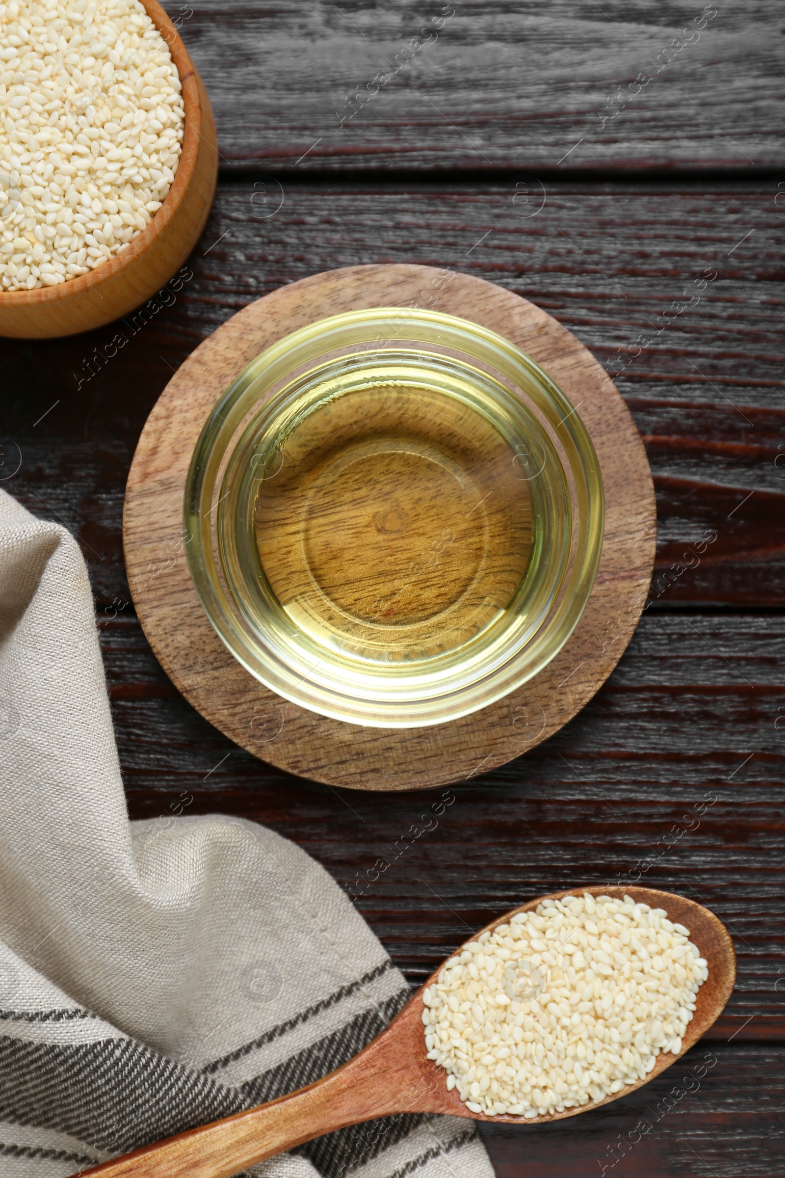 Photo of Fresh sesame oil in glass bowl and seeds on dark wooden table, flat lay