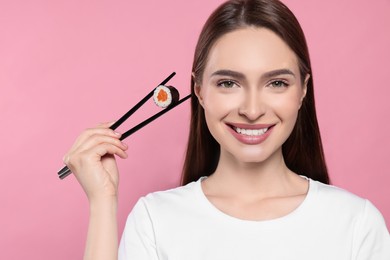 Beautiful young woman holding sushi roll with chopsticks on pink background