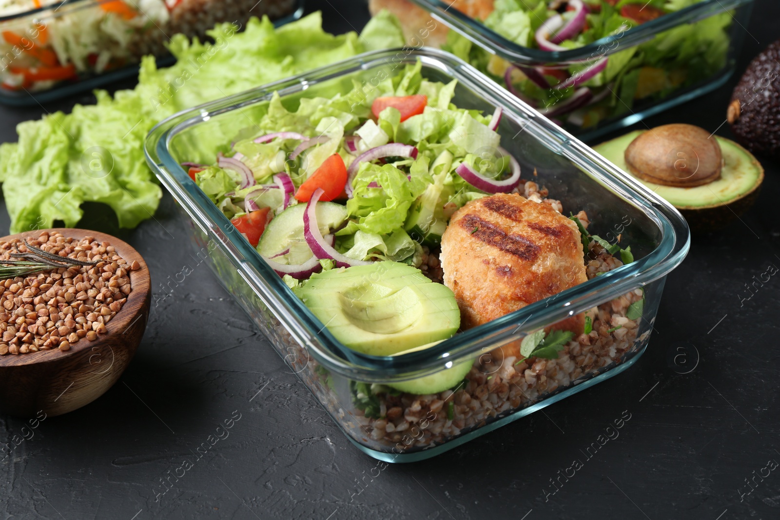 Photo of Healthy meal. Fresh salad, avocado, cutlet and buckwheat in glass container near other products on black table