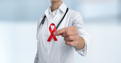 Doctor holding red awareness ribbon on blurred background, closeup. World AIDS disease day