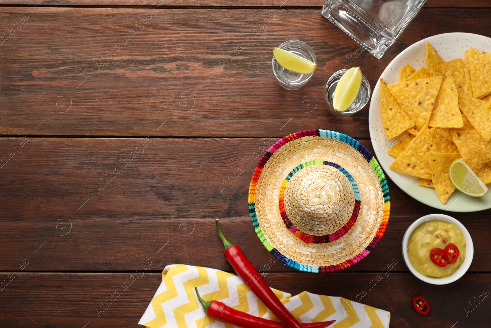 Photo of Mexican sombrero hat, tequila with lime, chili peppers, nachos chips and sauce on wooden table, flat lay. Space for text