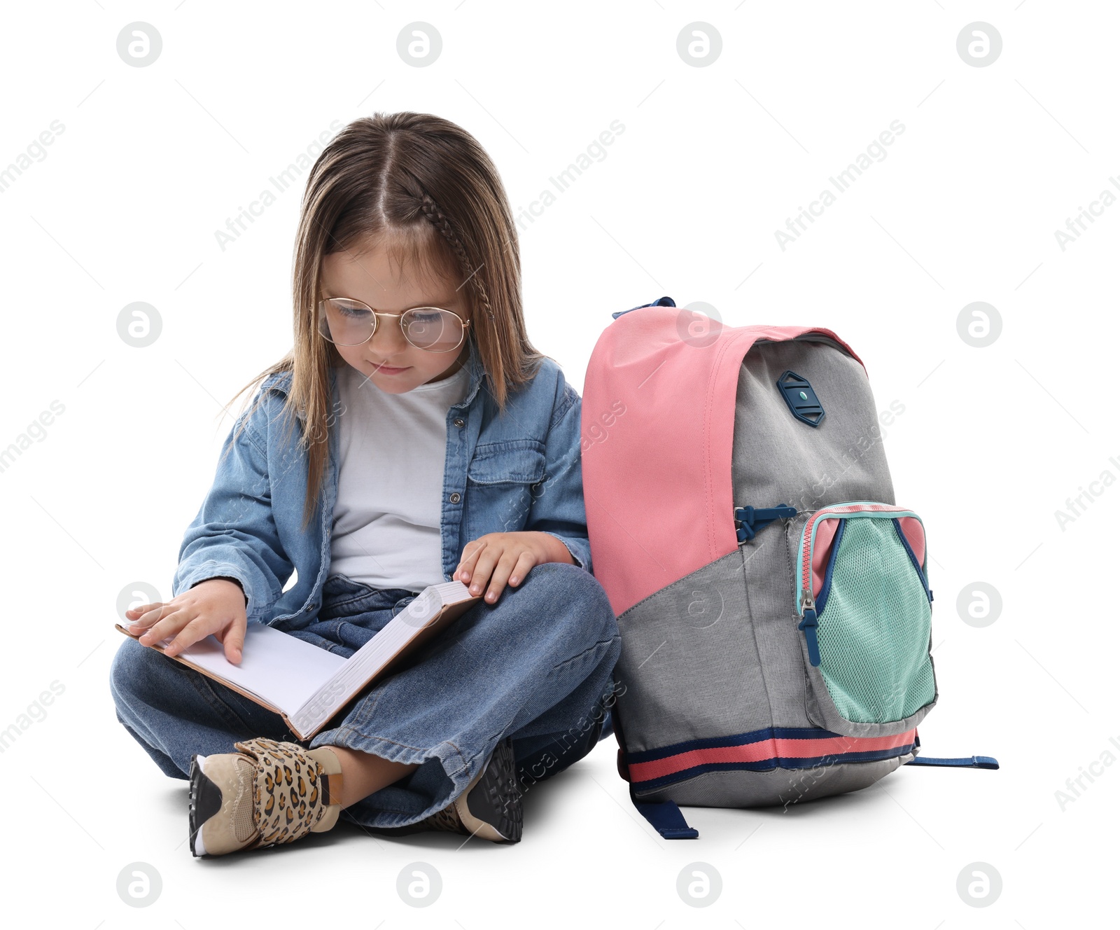 Photo of Cute little girl reading book near backpack on white background