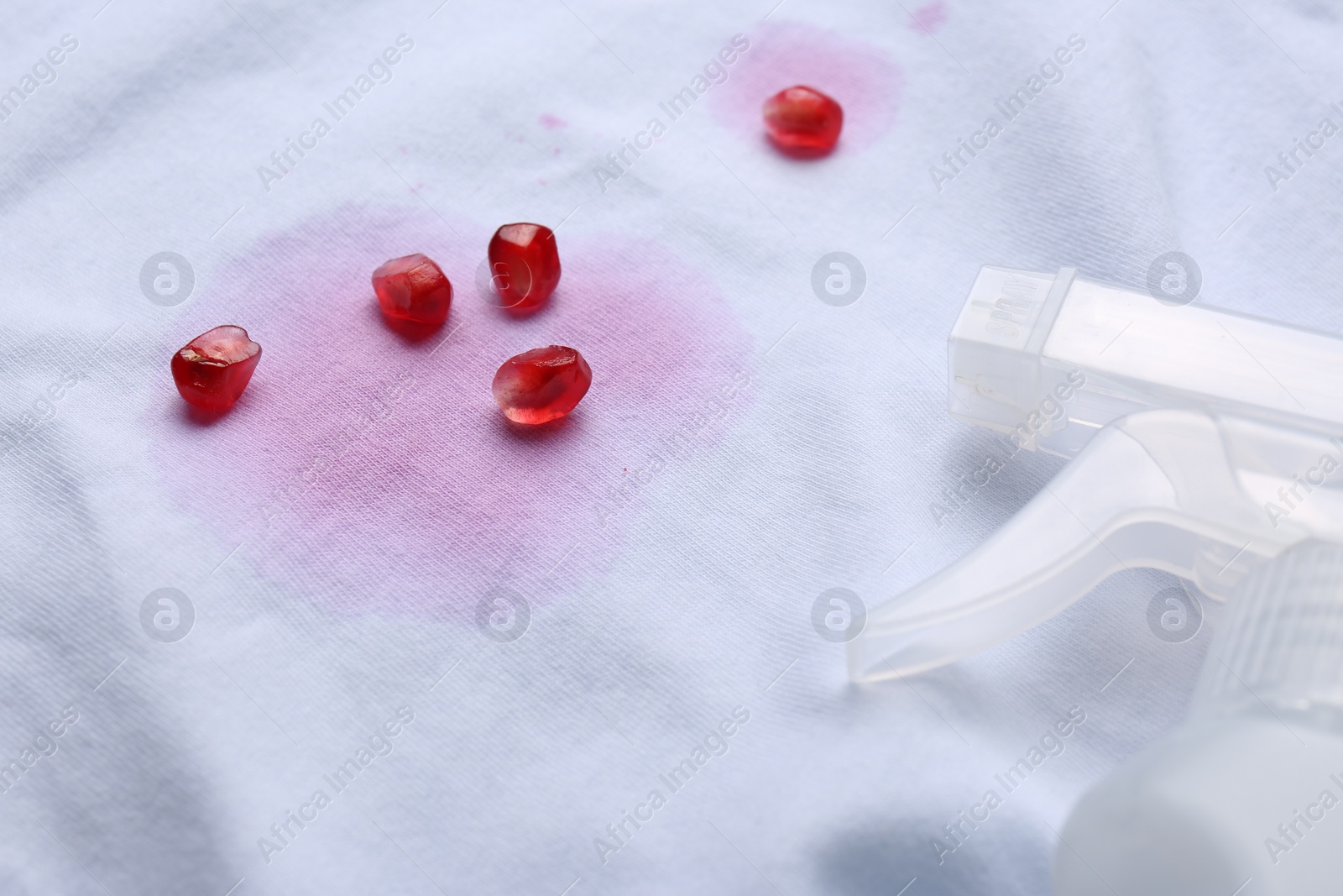 Photo of White shirt with fruit juicy stains, detergent and pomegranate seeds as background, closeup