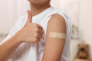 Photo of Boy with sticking plaster on arm after vaccination showing thumbs up indoors, closeup