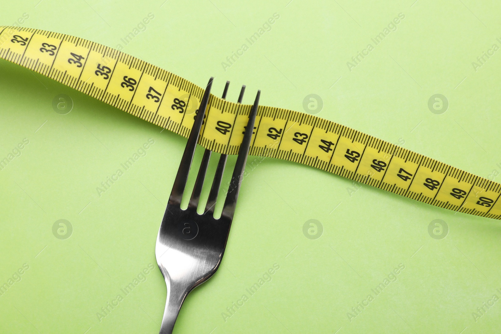 Photo of Fork with measuring tape on light green background, top view. Diet concept