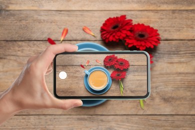 Woman taking picture of aromatic coffee and red gerbera flowers on wooden table, top view