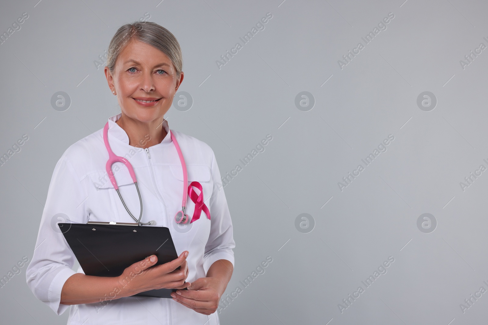 Photo of Doctor with pink ribbon, clipboard and stethoscope on light grey background, space for text. Breast cancer awareness