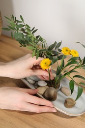 Photo of Woman creating stylish ikebana with beautiful flowers and green branches at wooden table, closeup