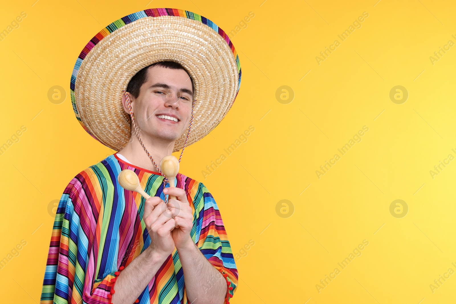 Photo of Young man in Mexican sombrero hat and poncho with maracas on yellow background. Space for text