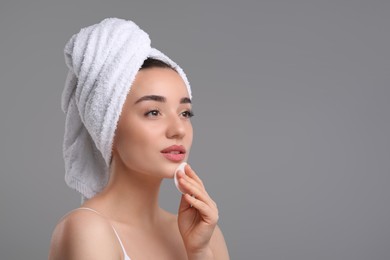 Beautiful woman in terry towel removing makeup with cotton pad on gray background, space for text