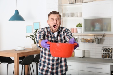 Photo of Emotional young man holding plastic basin under water leakage from ceiling in kitchen. Plumber service
