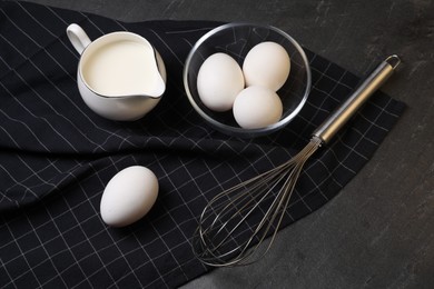 Photo of Metal whisk, raw eggs and milk on dark grey table