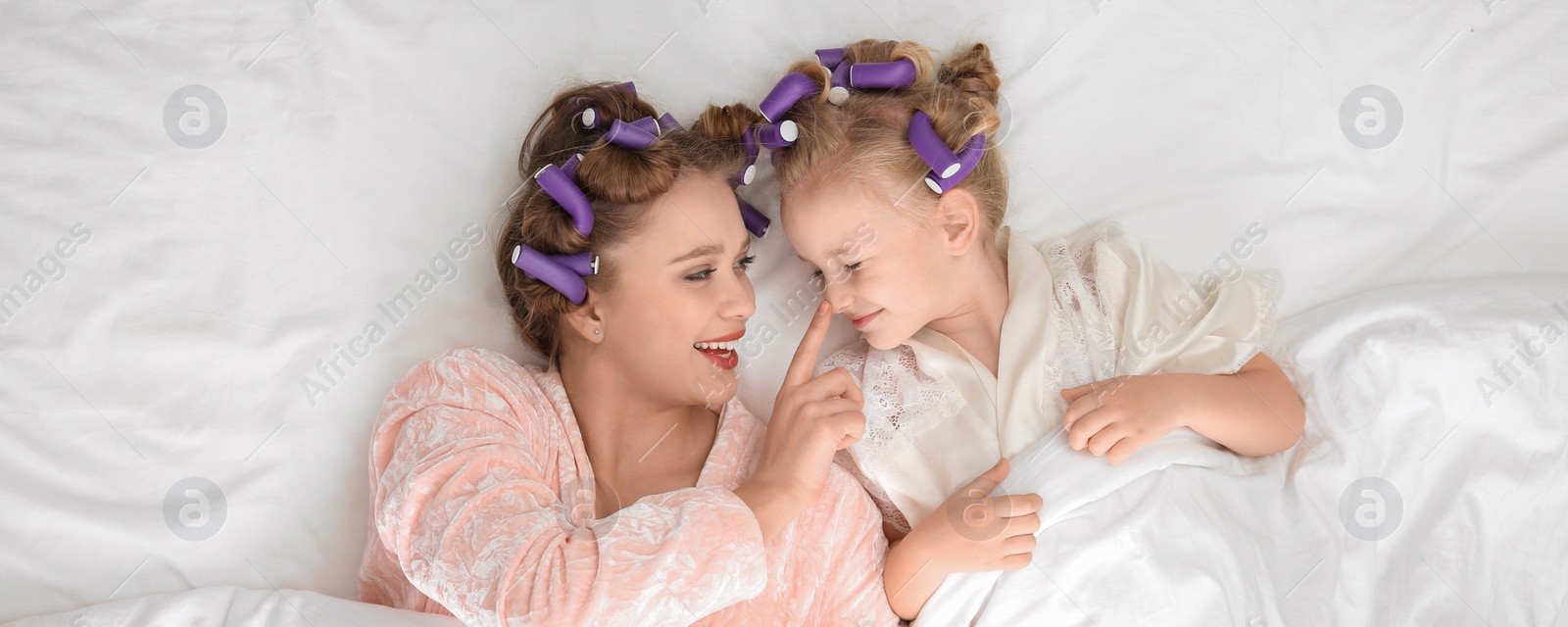 Image of Happy mother and daughter with curlers lying on bed, top view. Banner design