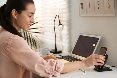 Photo of Woman putting mobile phone onto wireless charger at white table indoors. Modern workplace accessory