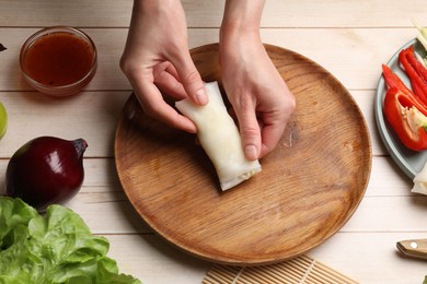 Photo of Making delicious spring rolls. Woman wrapping fresh vegetables into rice paper at wooden table, closeup