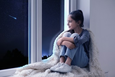 Image of Cute little girl sitting near window and looking at shooting star in beautiful night sky