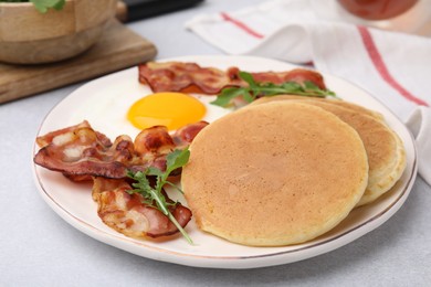 Photo of Plate with tasty pancakes, fried egg, arugula and bacon on light grey textured table, closeup