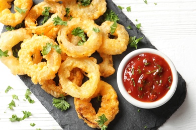 Photo of Homemade crunchy fried onion rings with tomato sauce on wooden table, top view