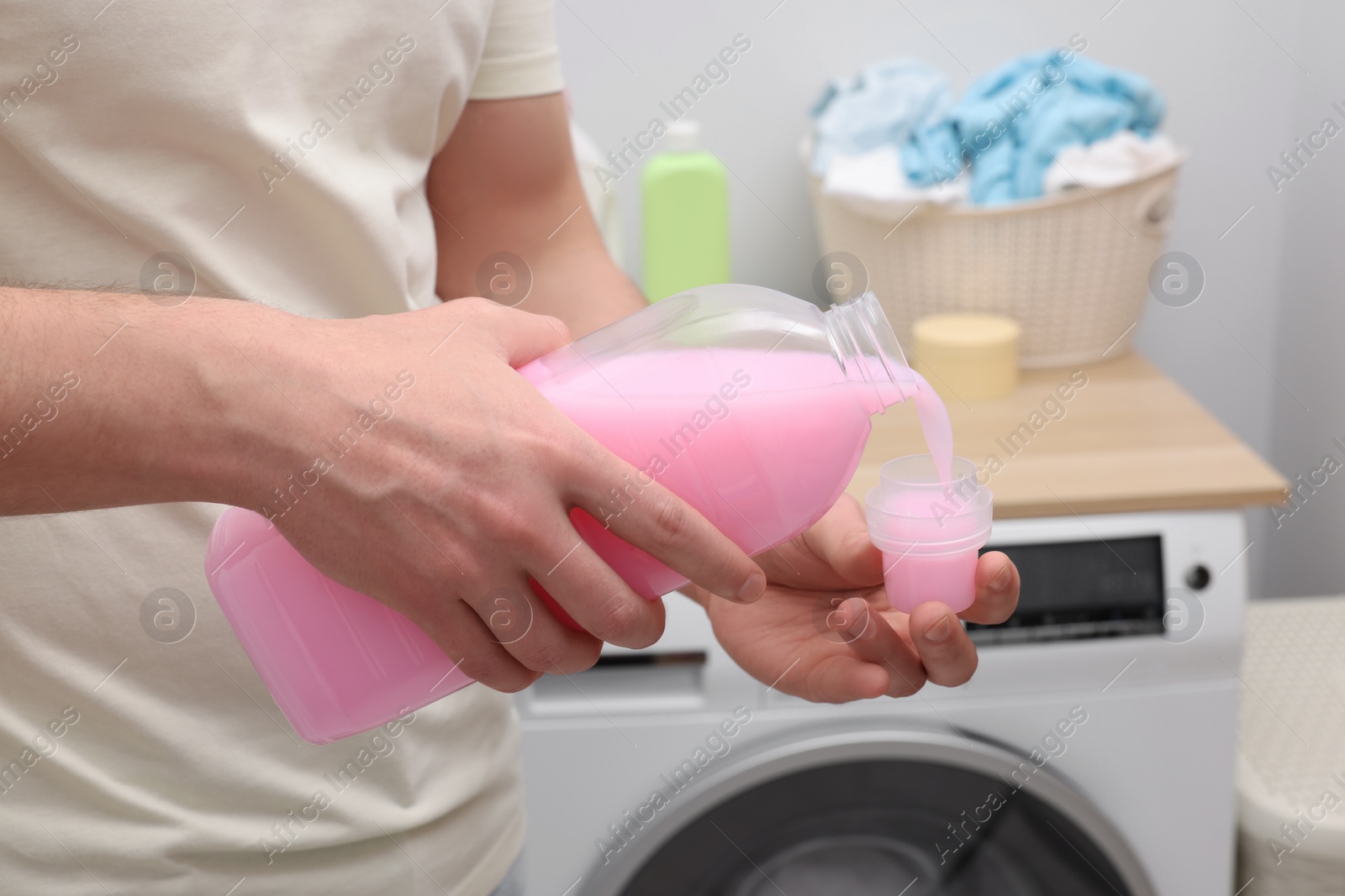 Photo of Man pouring fabric softener from bottle into cap near washing machine indoors, closeup