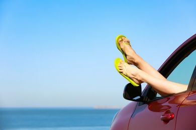 Photo of Closeup of woman showing legs with flip flops from car near sea, space for text. Beach accessories