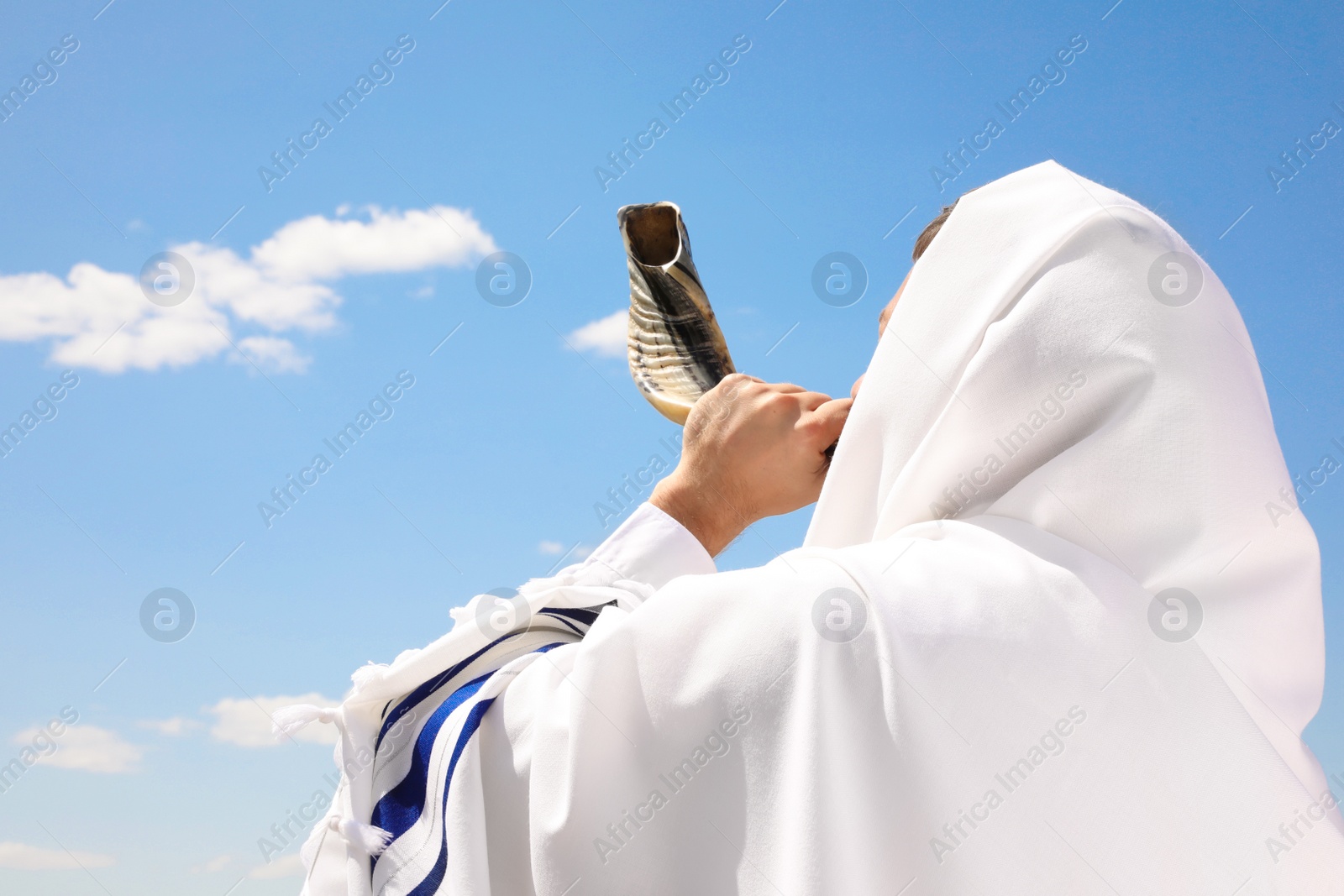 Photo of Jewish man in tallit blowing shofar outdoors. Rosh Hashanah celebration