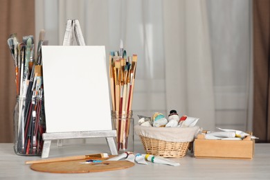 Photo of Many brushes, easel with blank canvas and paints on white wooden table indoors