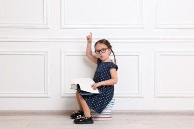 Photo of Cute little girl in glasses sitting on stack of books near white wall