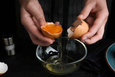 Photo of Woman separating egg yolk from white over glass bowl at black table, closeup